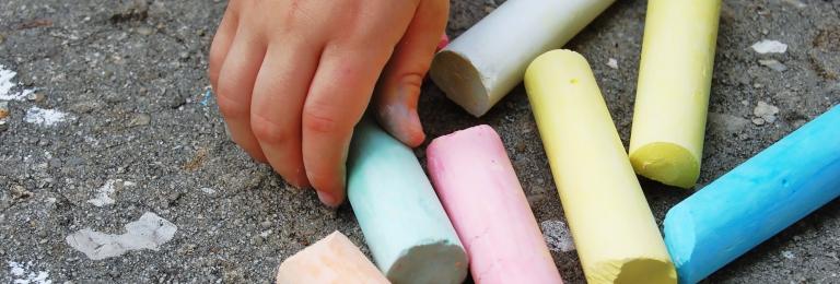 colourful chalk resting on the pavement. A child's hand reaching for one piece of chalk.